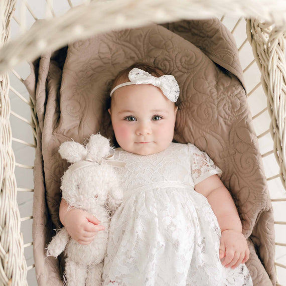 A baby wearing the Rose Romper Dress and matching Rose Bow Headband lies in a wicker bassinet with the Rose Bunny plush toy at her side, looking directly at the camera.