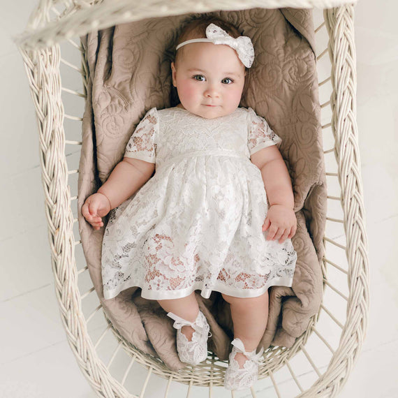 A baby dressed in the Rose Romper Dress and a matching Rose Bow Headband, lies comfortably in a woven basket, looking up towards the viewer with a gentle expression.