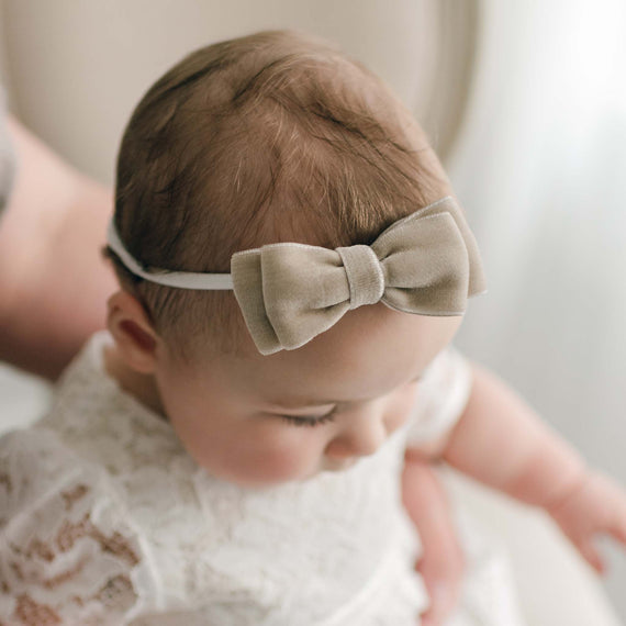 Close-up of a baby with a Rose Velvet Bow Headband and matching Rose Christening Gown, viewed from above, with a soft-focus background. The Rose Velvet Headband is a hand-stretched nylon band with a soft double face velvet ribbon tied in a bow.
