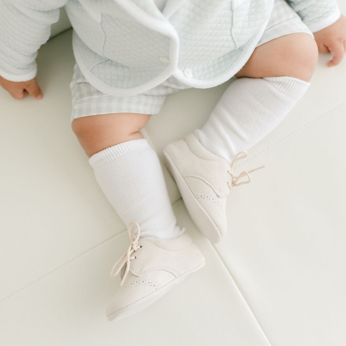A close-up of a baby's legs dressed in a designer pale blue quilted onesie and white socks, wearing Ian Suede Shoes, laying on a white surface.