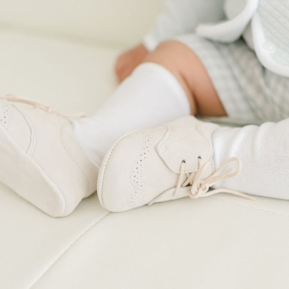 Close-up of a baby's feet wearing white Ian Suede Shoes and white leggings, resting on a light beige background. The focus is on the delicate shoes and leggings.