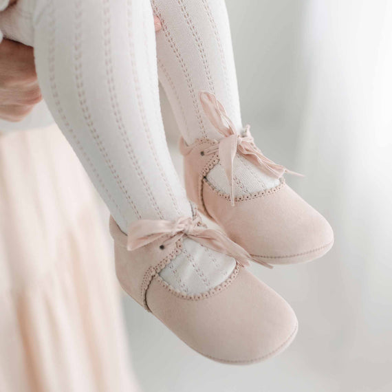A close-up of a baby's feet in Joli Suede Tie Mary Janes adorned with delicate bows, complemented by a pair of white knit leggings. The backdrop is soft and blurred.