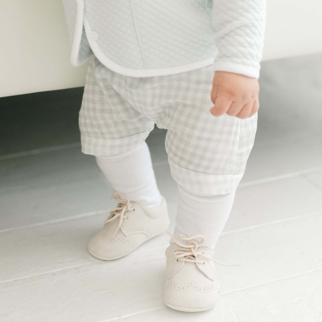 Close-up of a toddler's lower half wearing beige designer shoes, white socks, and Ian Cloud Shorts Set under a light blue quilted jacket, highlighting small, cautious steps.
