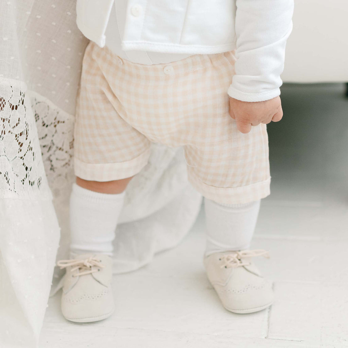 A toddler in traditional beige checkered shorts and Ian Suede Shoes stands next to a lace curtain, focusing on their tiny feet and hands.