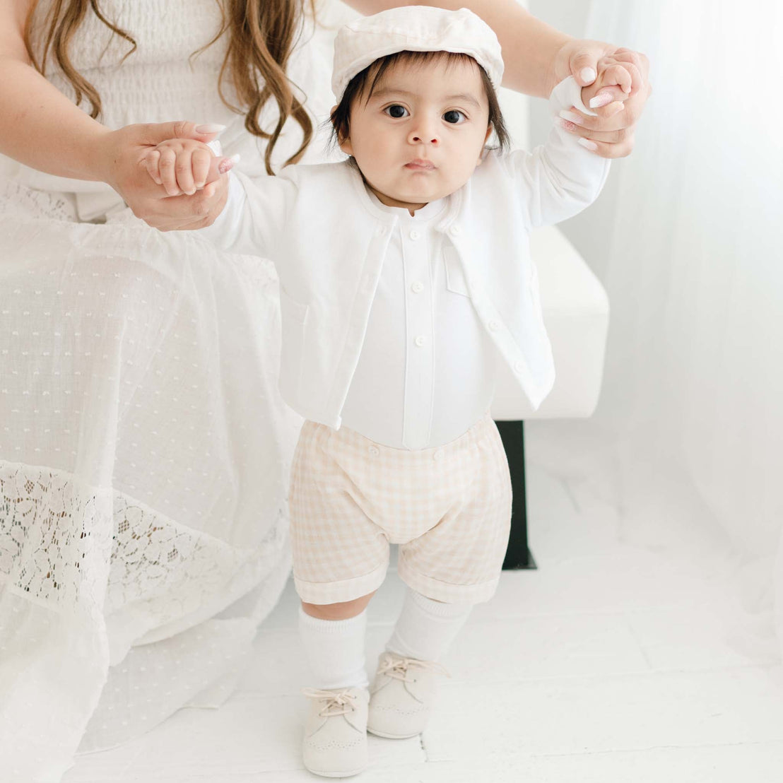 A baby wearing an Ian Fawn Shorts Set with traditional plaid shorts and a cute bonnet stands supported by a seated woman in a white dress, only her arms visible, in a bright room.