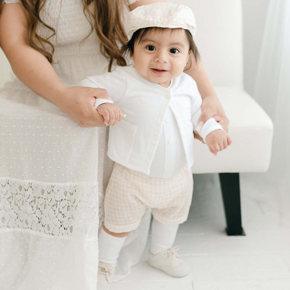 A baby taking steps with support, dressed in an Ian Fawn Shorts Set, holding hands with a woman in a designer white dress, in a light, airy room.