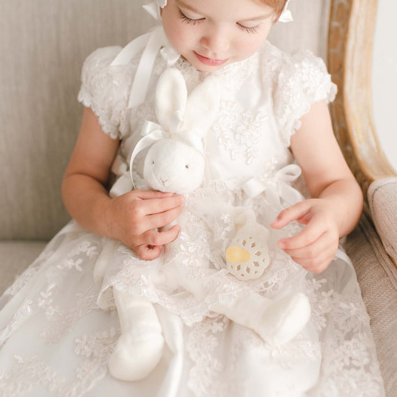 Baby girl holding plush bunny toy wearing christening dress.