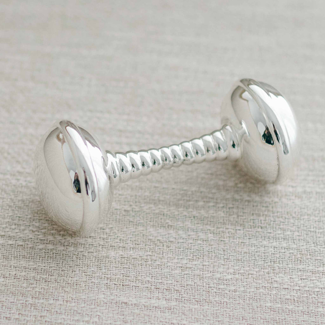A luxury vintage Silver Baby Rattle with two rounded ends and a twisted handle, lying on a textured beige fabric.