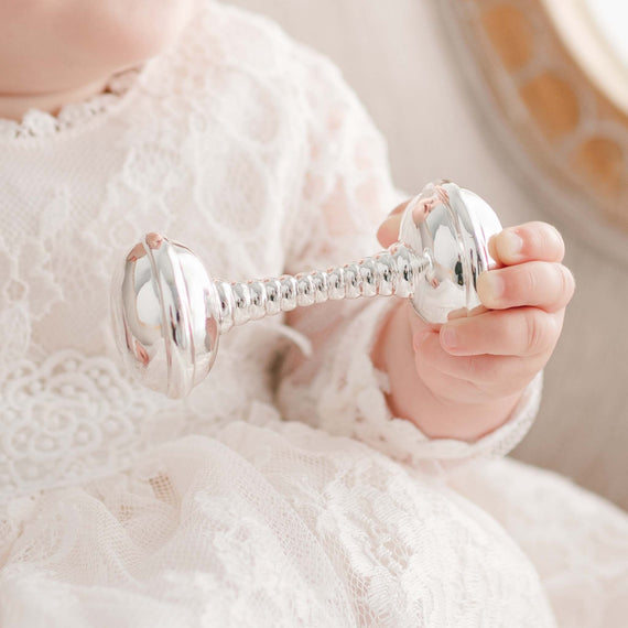 A close-up of a baby’s hand holding a Silver Baby Rattle, dressed in a white lace outfit, with a soft-focus background.