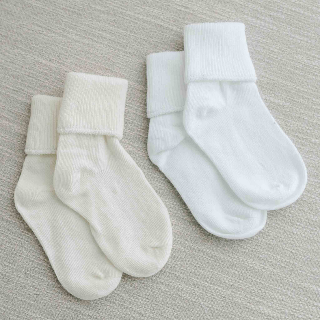 Two pairs of Boys Simple Socks, one pair in ivory and another in white, neatly laid out on a soft beige fabric background.