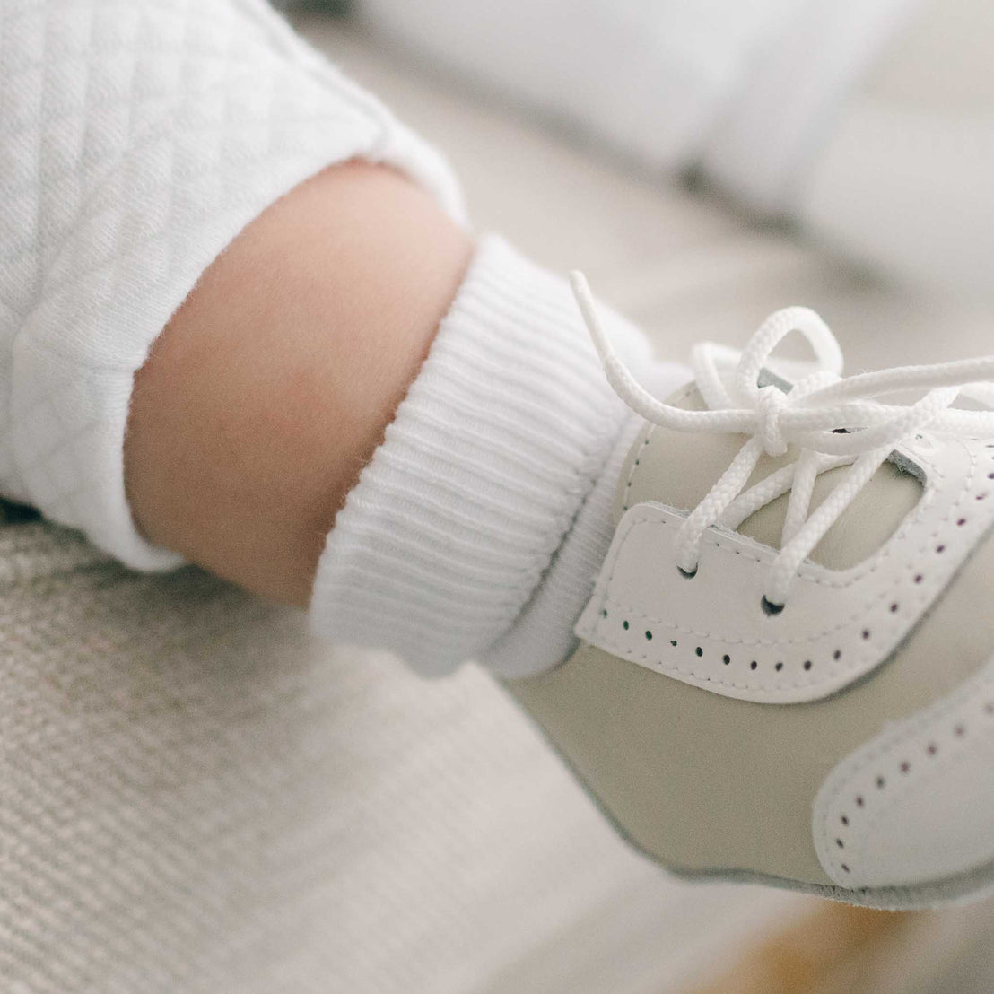 Close-up of a baby's foot clad in a Boys Simple Socks in white and the Boys Ivory Two Tone Wingtip Shoes.