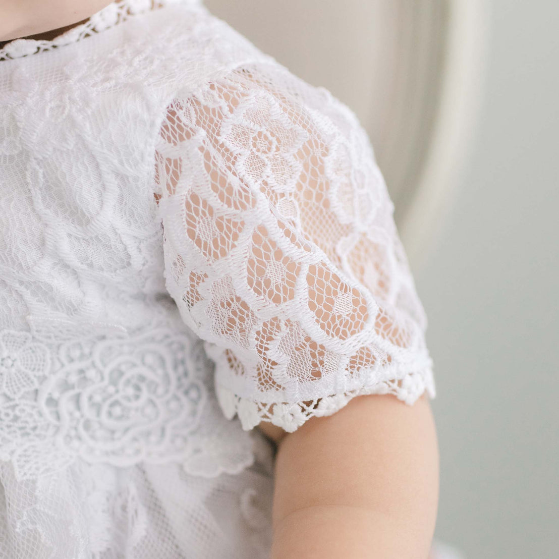 Close-up of a child's shoulder wearing the Olivia Christening Gown & Bonnet, highlighting the intricate floral pattern and scalloped edges of the fabric for a christening.