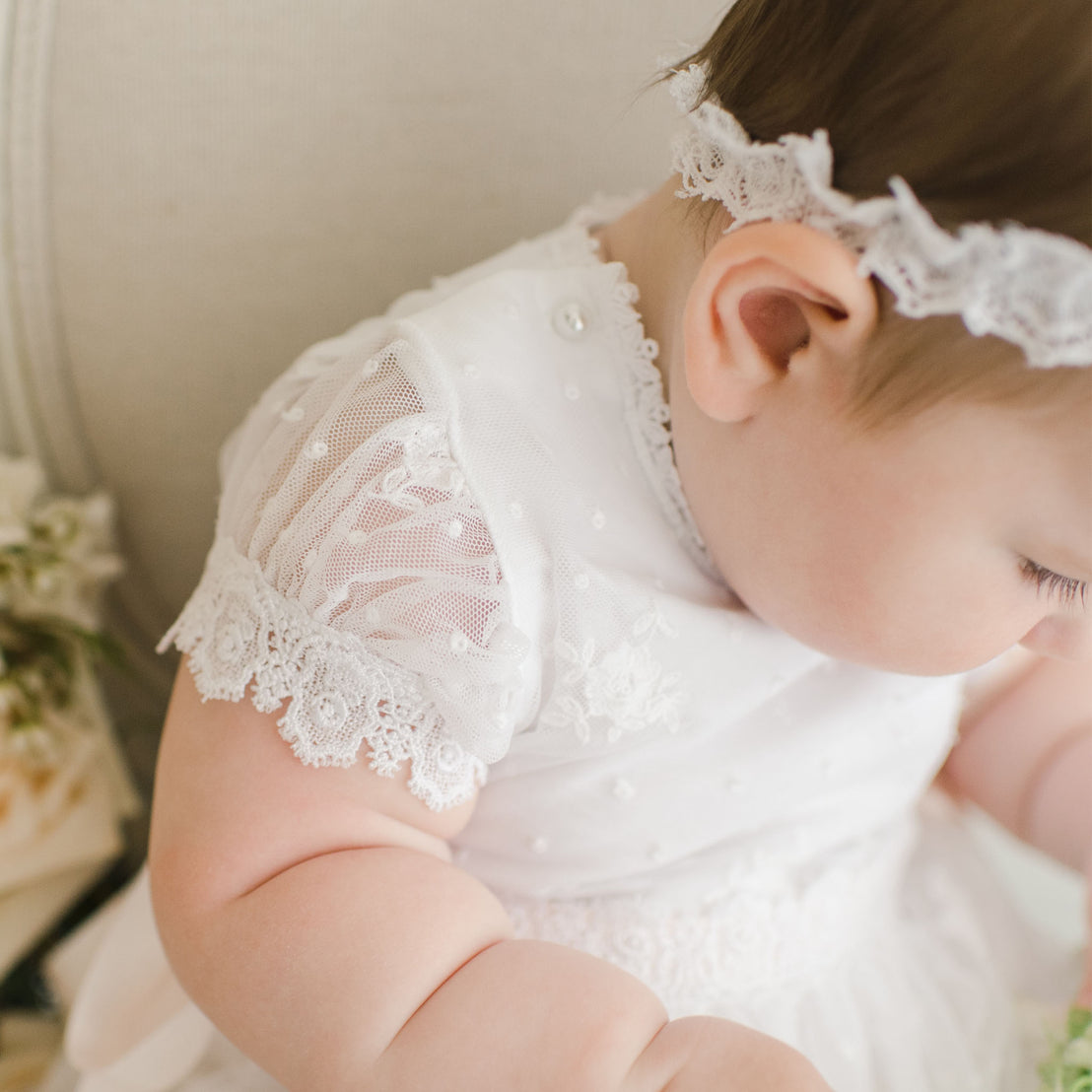 A close-up of a baby wearing the Melissa Romper Dress, a delicate white lace dress with intricate details, focused on the sleeve and part of the back. The baby's gaze is directed downwards.