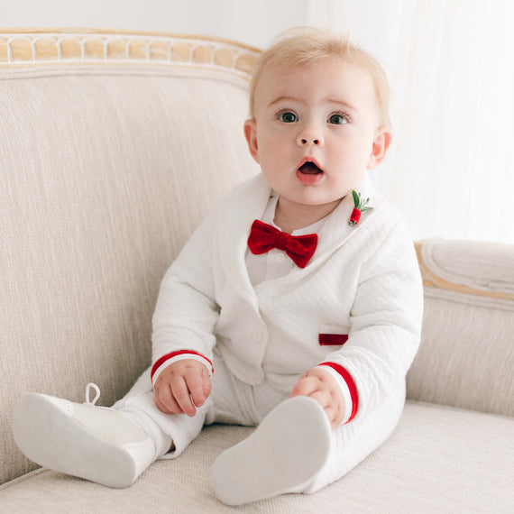 A baby with blue eyes and light hair wearing a Noah 3-Piece Suit with Red Trim sits on an upscale beige sofa, looking surprised.