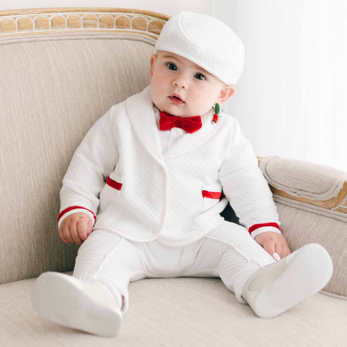 A baby dressed in a white christening outfit with red accents, including a Noah Red Bow Tie & Boutonniere, sitting on a beige sofa and looking to the side.