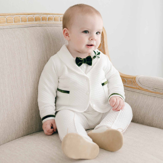 A baby wearing an elegant, upscale Noah 3-Piece Suit | Green Trim with a bow tie sits on a beige sofa, looking slightly to the side with a curious expression.