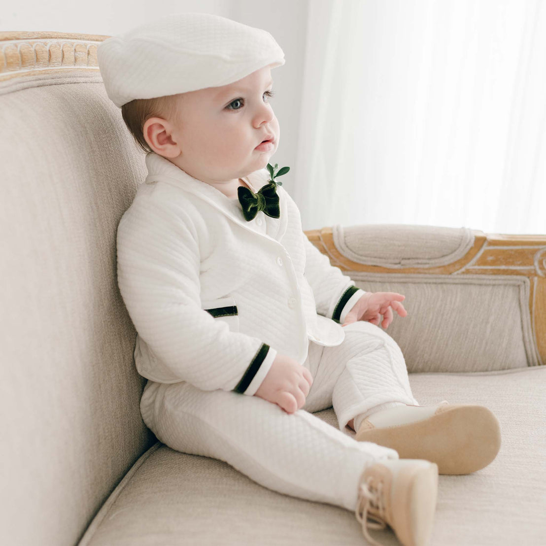A baby dressed in a Noah 3-Piece Suit | Green Trim with a hat and bow tie, sitting pensively on a cream sofa in a bright room for a christening ceremony.