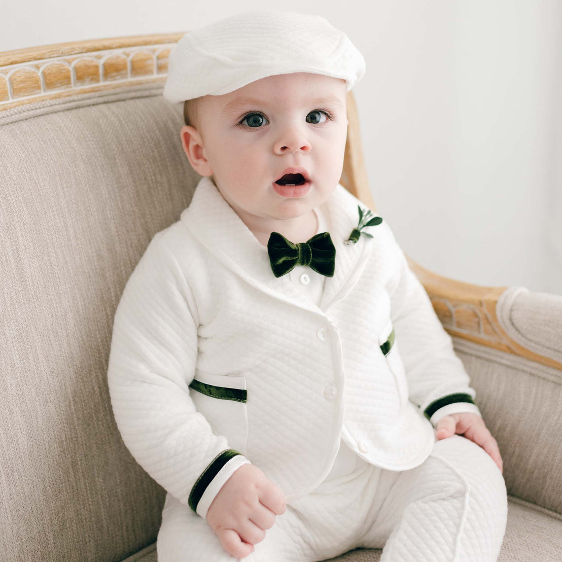 A baby wearing a Noah 3-Piece Suit with Green Trim and a cap sits on an upscale beige sofa, looking surprised with wide eyes.