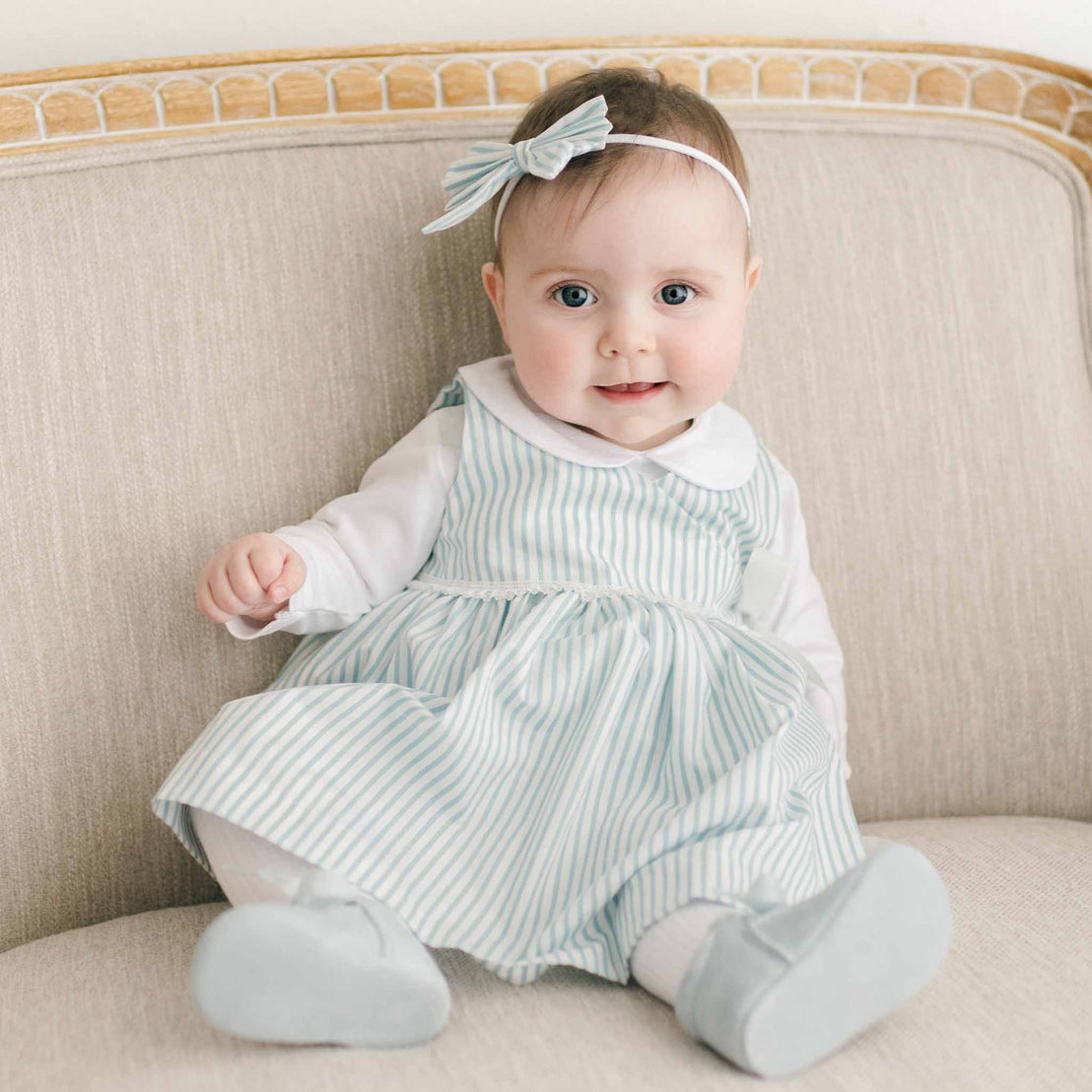 A baby in the blue Thea Wrap Dress and matching Thea Bow Headband sits on a beige sofa, looking curiously at the camera.