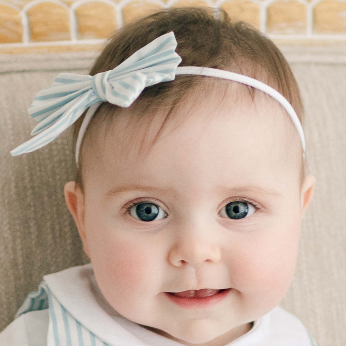 A close-up of a baby with bright blue eyes and a small smile, wearing the blue Thea Bow Headband against a beige fabric background.