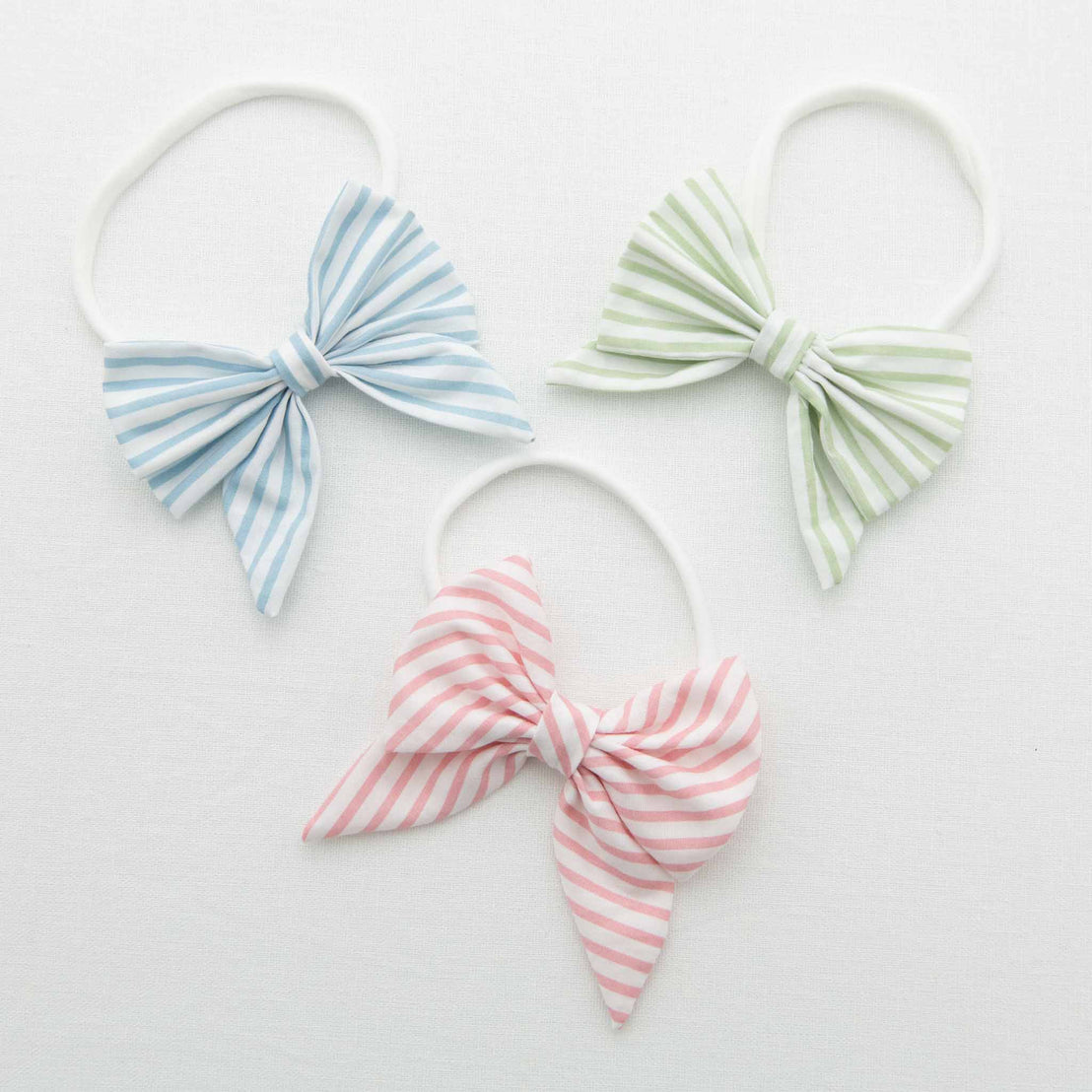 The three shades of the Thea bow headbands in blue, green, and pink on a white background.
