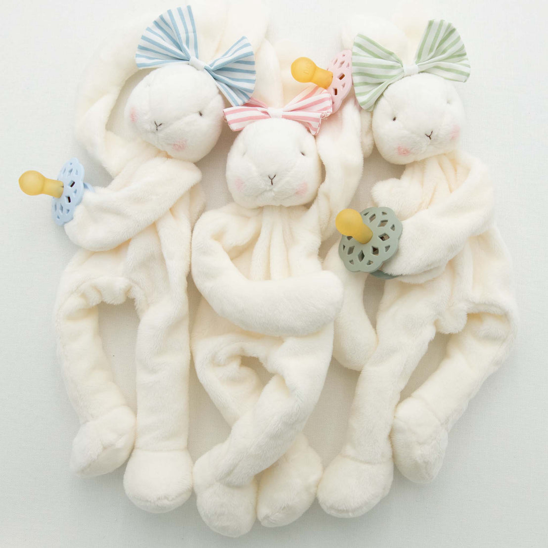 Three Thea Bunny Buddy with pacifiers on a white background, each featuring a different colored miniature version of the Thea Bow Headband: blue, pink, and green.