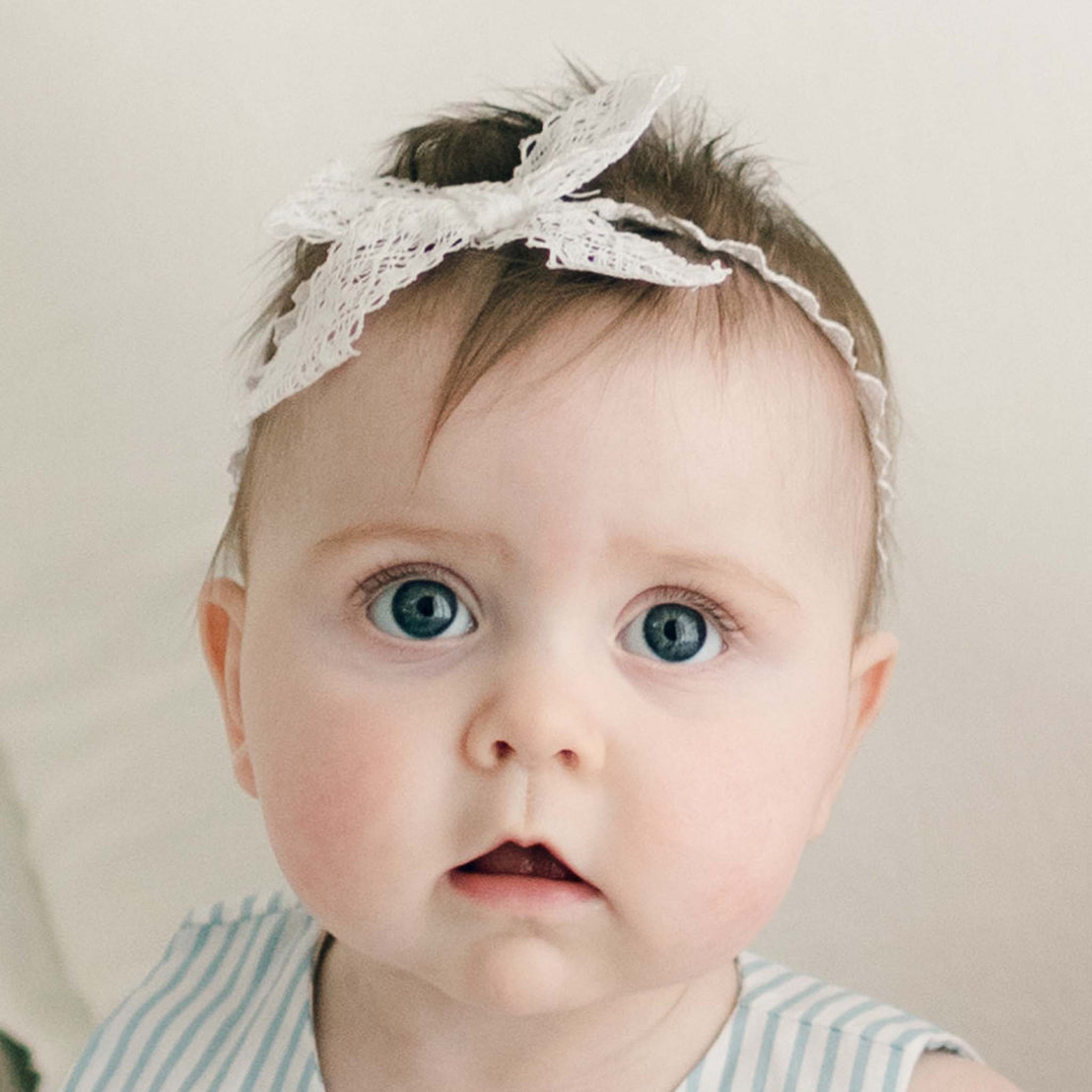 A close-up portrait of a baby with large blue eyes and dark hair, wearing the Thea Lace Headband, dressed the blue Thea Wrap Dress, against a neutral background.