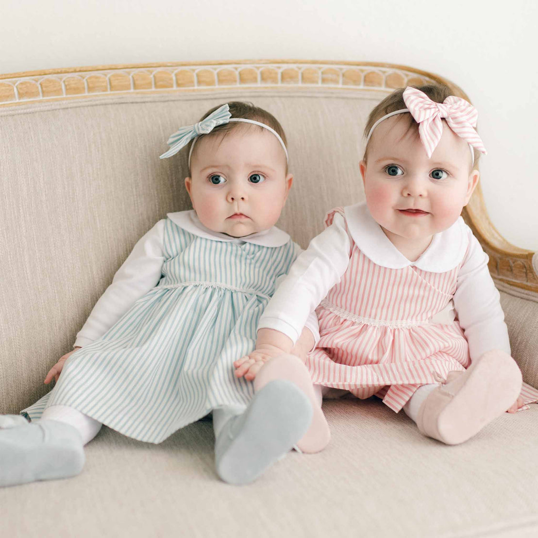 Two twin baby girls in the Thea Wrap Dresses, with white linen collar onesie underneath, and matching Thea Bow Headband sitting on a beige sofa, one looking curious and the other smiling at the camera.