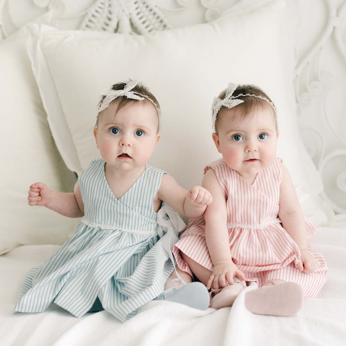 Two twin baby girls wearing the Thea Wrap Dress, one in blue and the other in pink, and matching Thea Lace Headband sit on a white bed, surrounded by pillows, looking directly at the camera.