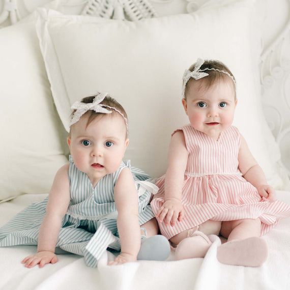 Two twin baby girls dressed in the Thea Wrap Dresses, one in blue and the other in pink, and matching Thea Lace Headbands, sitting on a white couch with pillows, looking curiously towards the camera.