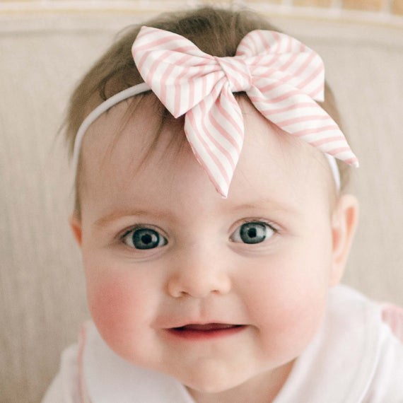 A baby with bright blue eyes and rosy cheeks smiles at the camera, wearing the pink Thea Bow Headband.