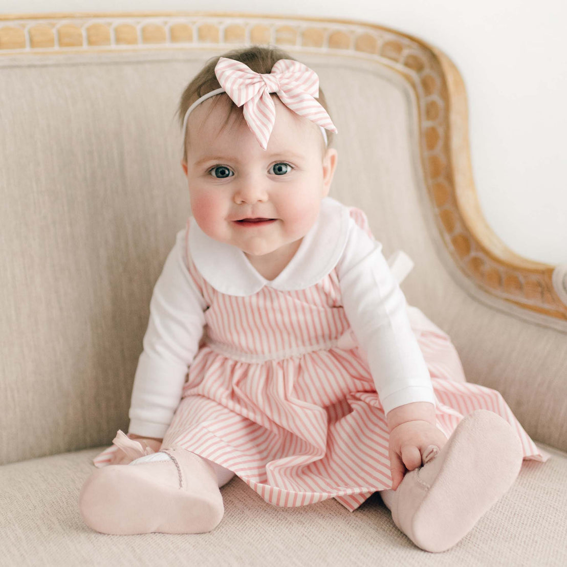 A cheerful baby with bright blue eyes in a Thea Wrap Dress, a striped pink and white dress, with the matching Thea Bow Headband, sitting on a beige sofa.