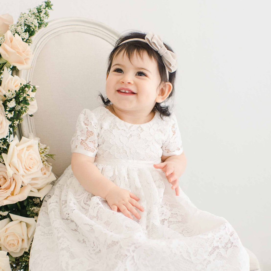A joyful toddler wearing a traditional white lace dress sits beside a chair adorned with pink roses, smiling brightly in a vintage, airy room while wearing the Rose Velvet Bow Headband.