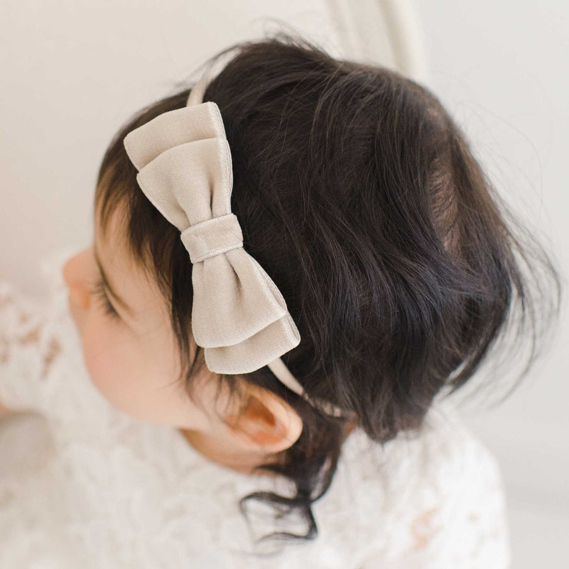 A close-up image of a baby wearing a Rose Velvet Bow Headband on her dark hair, focusing on the top and side of her head against a soft, light background.