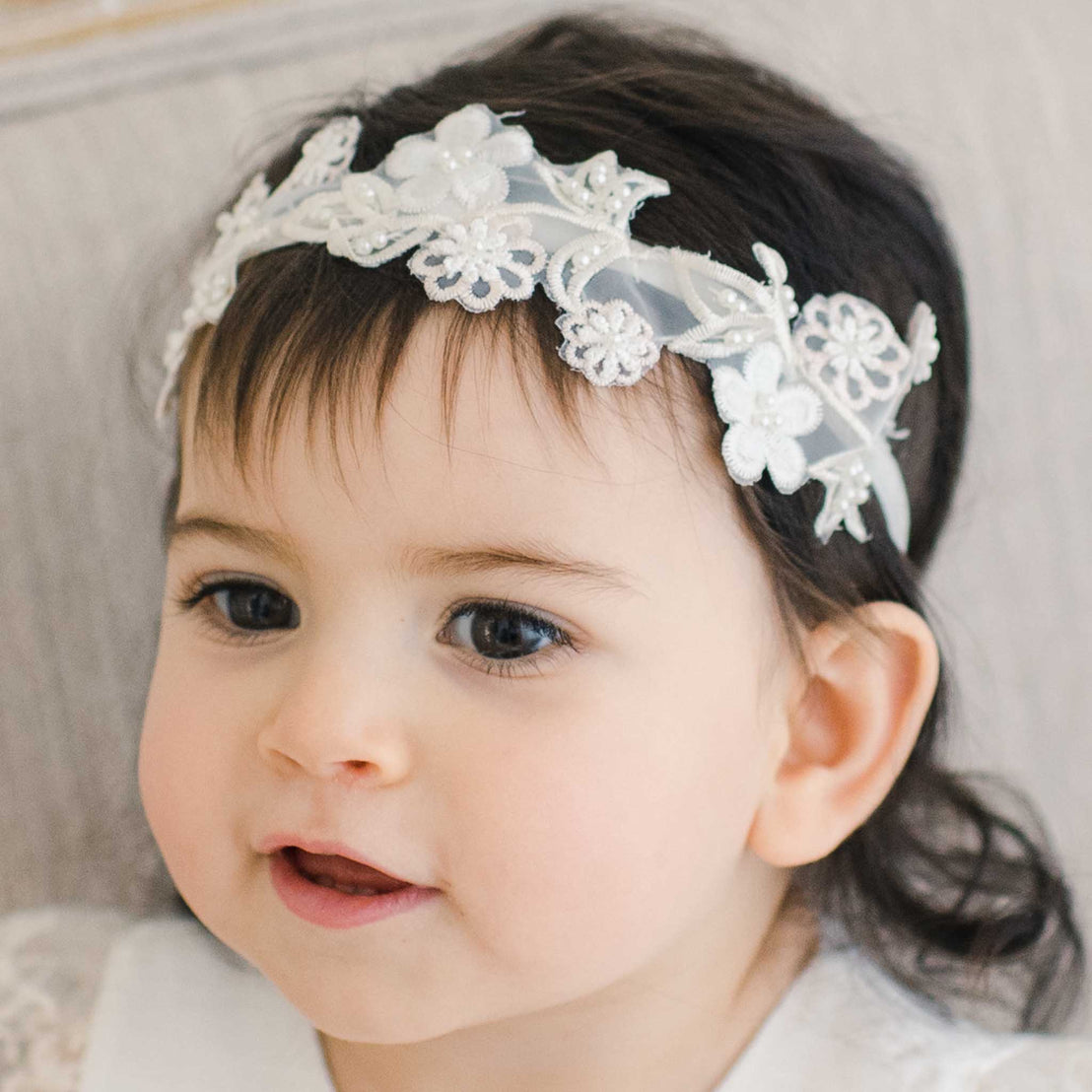 Close-up of a toddler with dark hair wearing the Jessica Beaded Flower Headband, a delicate white floral headband, showing a joyful expression.
