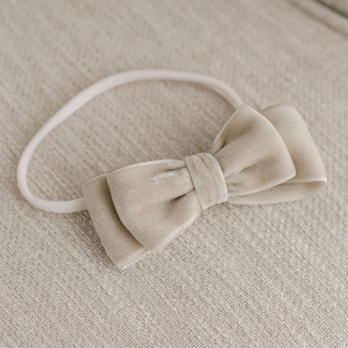 A rose velvet bow headband neatly placed on a textured grey fabric surface. The bow has a soft and luxurious appearance with a simple, elegant design, giving it an upscale, vintage look.