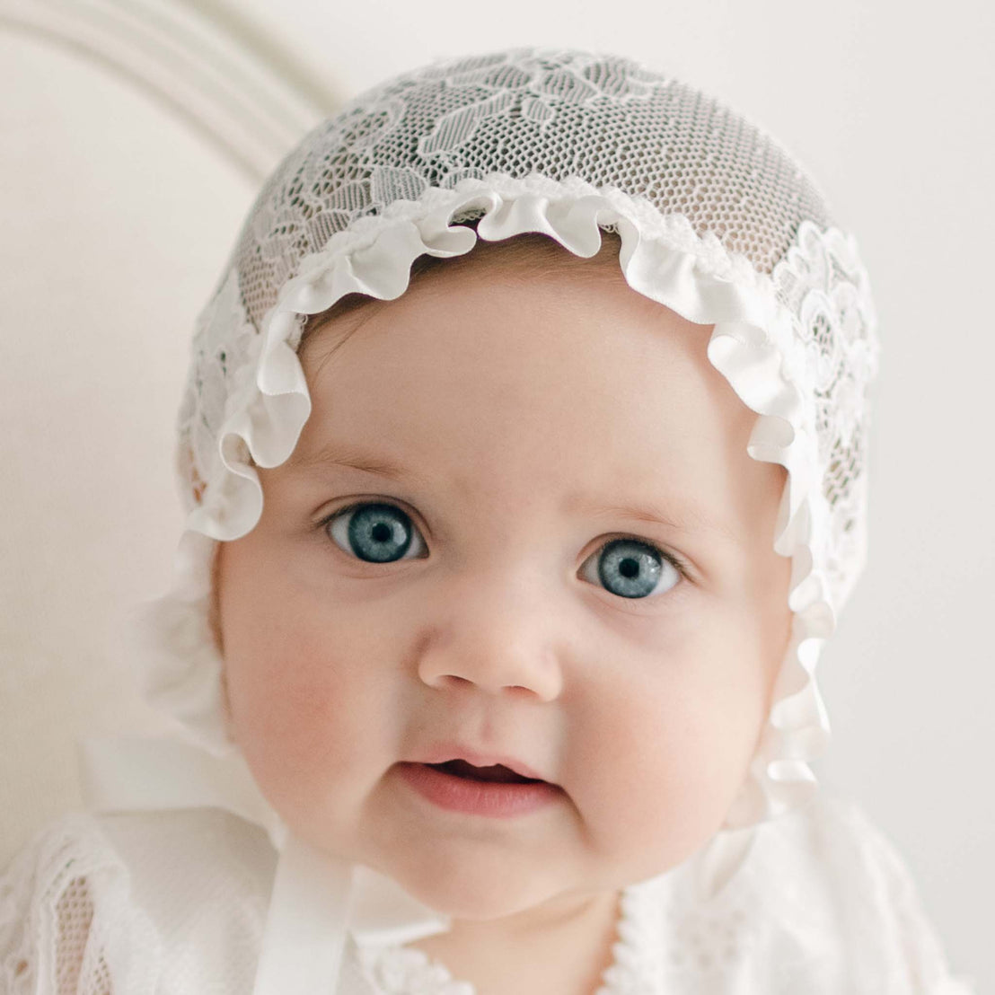 A baby girl with big blue eyes is wearing the Victoria Lace Bonnet with ruffled edges, complementing her Victoria Puff Sleeve Christening Gown. The baby looks directly at the camera with a smile. The background is softly lit and plain.