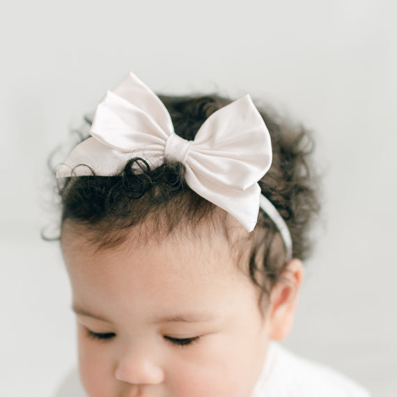 A close-up of a baby with curly hair wearing a Victoria Silk Bow Headband, set against a light gray background. The baby has a thoughtful expression.