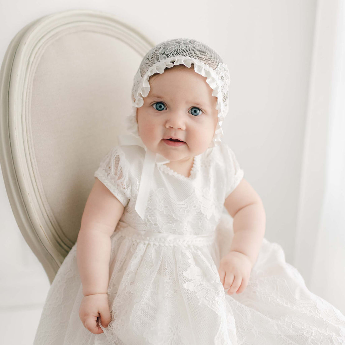 A baby wearing the Victoria Puff Sleeve Christening Gown and Bonnet sits on a light-colored, upholstered chair. The baby has blue eyes and a slight smile.