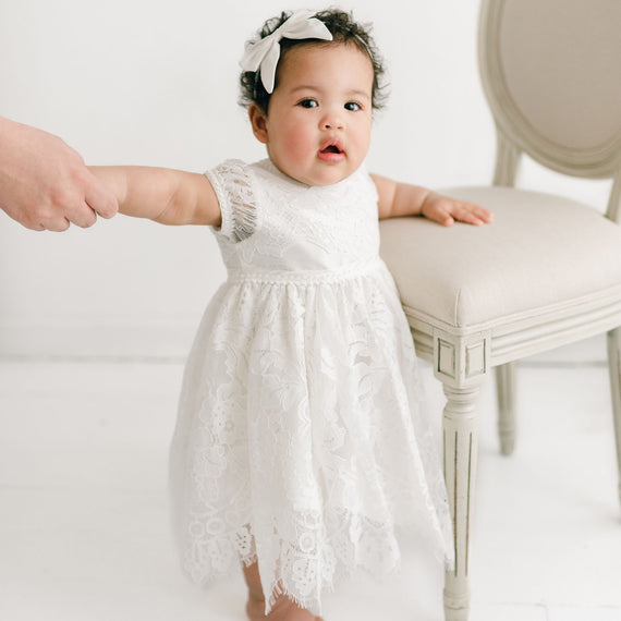 A toddler in a Victoria Puff Sleeve Christening Dress and headband stands by a chair, holding an adult's hand, in a light, elegant room.
