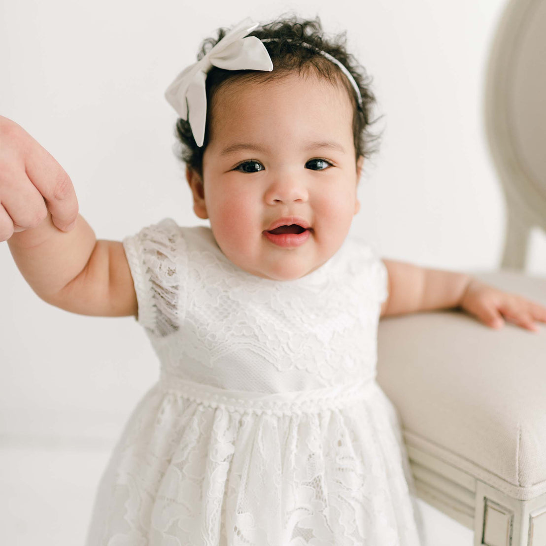 A cheerful baby girl with curly hair wearing a Victoria Puff Sleeve Christening Dress and a white bow headband smiles while reaching out her hand, standing next to a light-colored chair in a brightly lit room.
