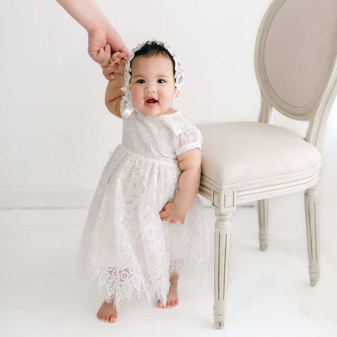 A baby girl in a Victoria Puff Sleeve Christening Dress standing beside a chair, holding an adult's hand. She wears a white headband and looks forward with a joyful expression.