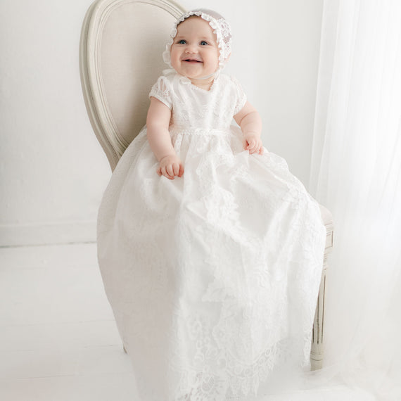 A smiling baby wearing a Victoria Puff Sleeve Christening Gown & Bonnet sits on a vintage, light-colored chair. The gown features ivory embroidered lace over a silk Dupioni lining. The The background is white and softly lit.