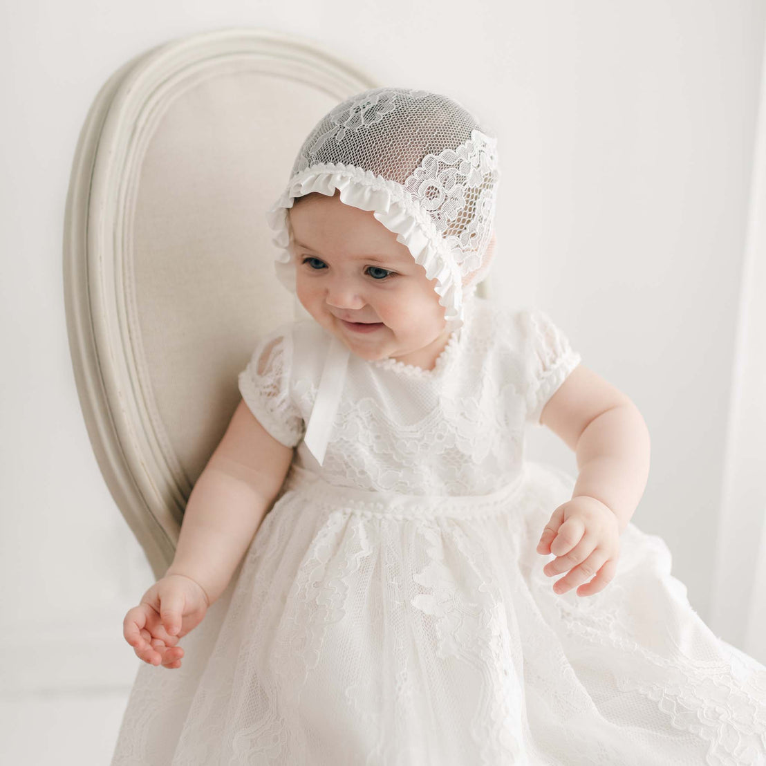 A baby wearing the Victoria Puff Sleeve Christening Gown and Bonnet sits on a cream-colored upholstered chair. The gown is made with embroidered ivory floral lace and silk Dupioni lining. The baby is smiling and looking to the side. The background is softly lit and white.