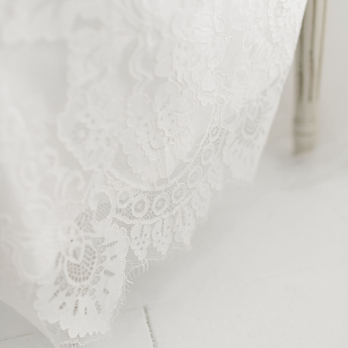 Close-up of delicate embroidered ivory floral lace featuring intricate floral patterns. The Victoria Puff Sleeve Christening Gown & Bonnet, with its elegant details clearly visible, drapes smoothly over a silk Dupioni lining, and a portion of a wooden chair leg appears in the background. The scene is well-lit and crisp.