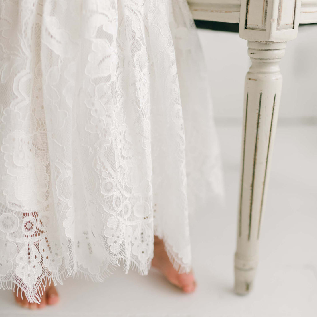 A close-up image of a person's bare feet peeking from under a delicate white Victoria Puff Sleeve Christening Dress, next to a vintage white wooden chair. The focus is on the intricate embroidered lace pattern and the texture of the chair.