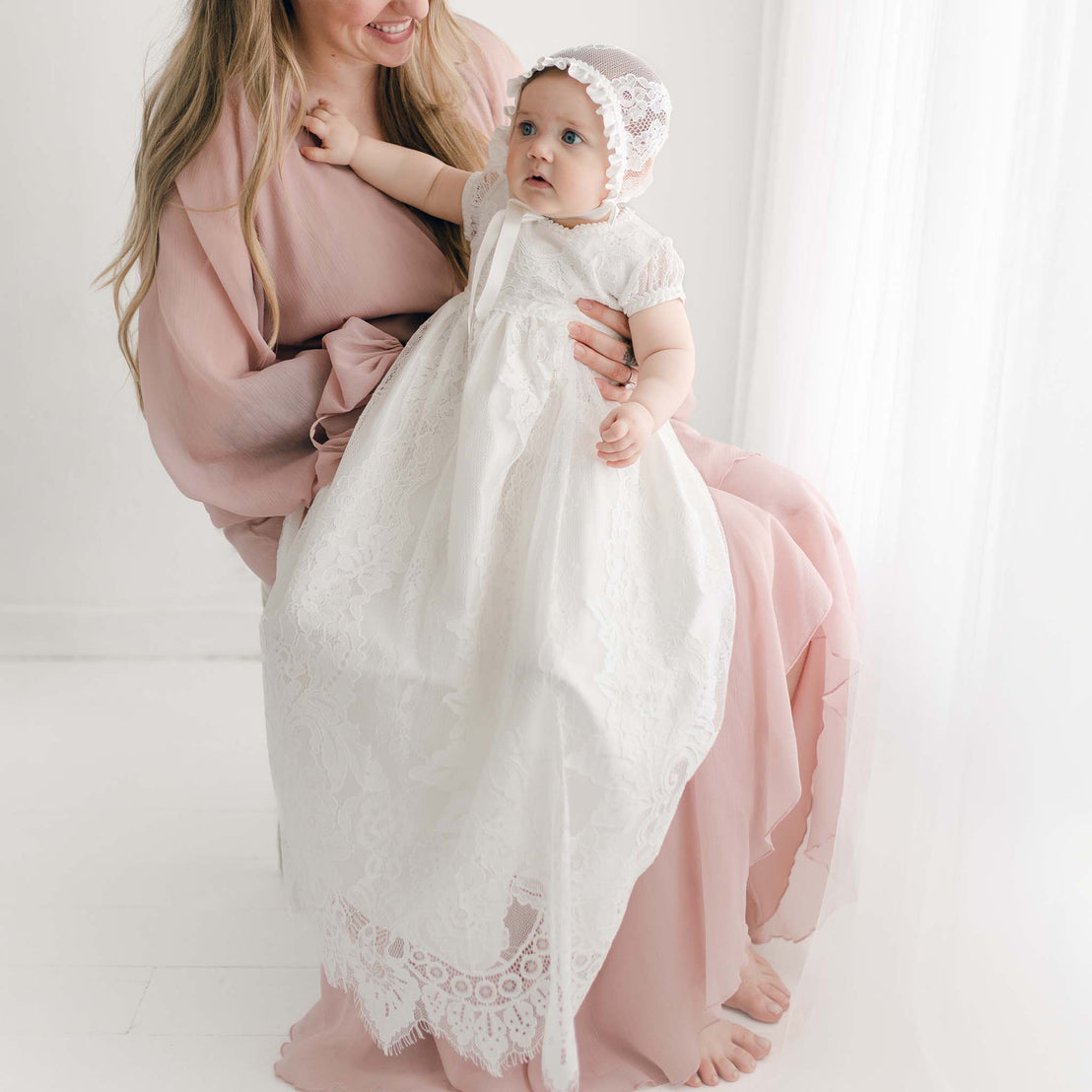 A woman in a light pink dress sits, holding a baby dressed in the Victoria Puff Sleeve Christening Gown and Bonnet. The baby looks curious, reaching up. The background is softly lit with a white curtain.