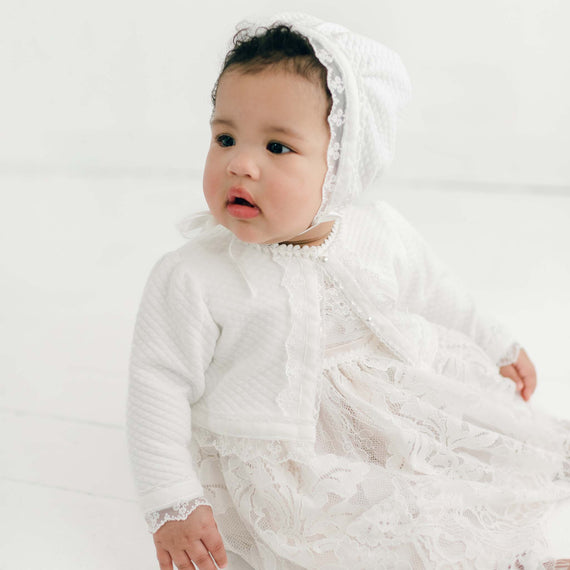 A baby dressed in a Victoria Quilted Cotton Sweater gown and bonnet looks off to the side with a curious expression in a bright, all-white studio setting.