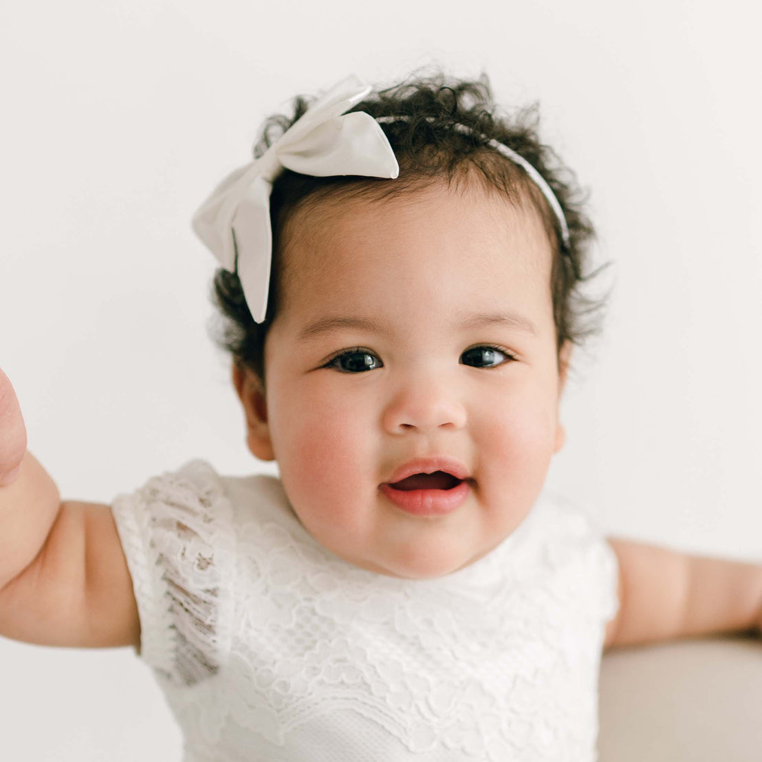 A baby girl with curly hair and a Victoria Silk Bow Headband smiles joyfully, wearing a lacey white dress against a clean, light background.