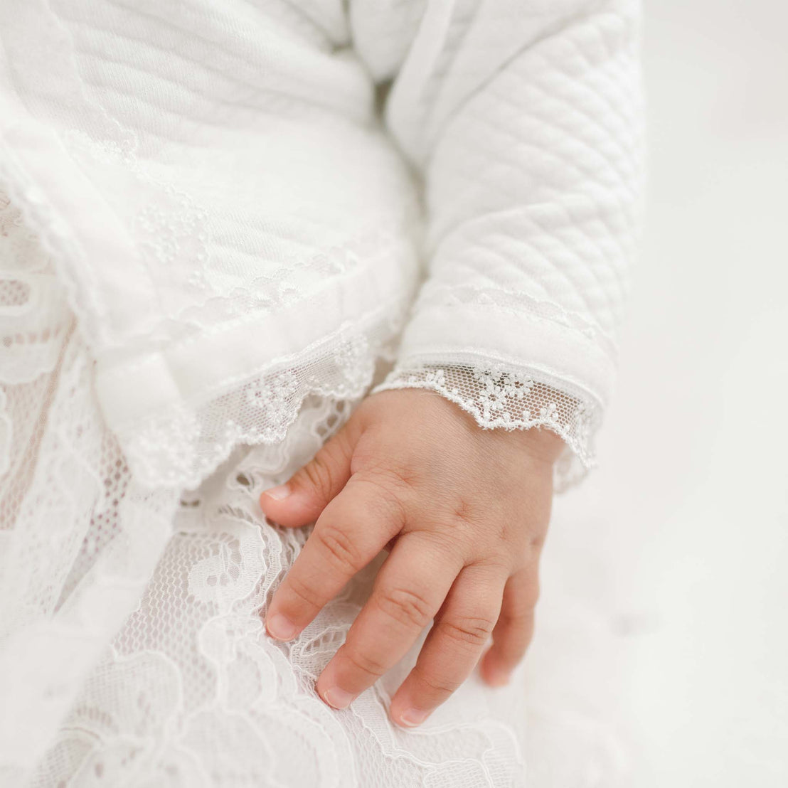 Close-up of a baby's hand gently resting on a Victoria Quilted Cotton Sweater, capturing the delicate, soft skin and tiny fingers.